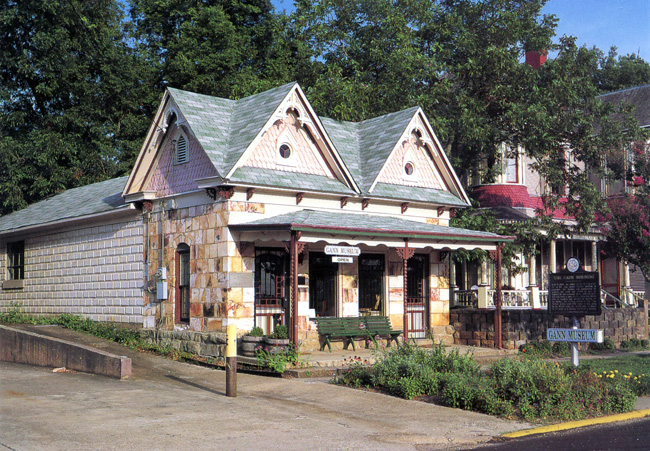 Stone building with sign "Gann Museum," wooden gables, covered porch, garden along street.