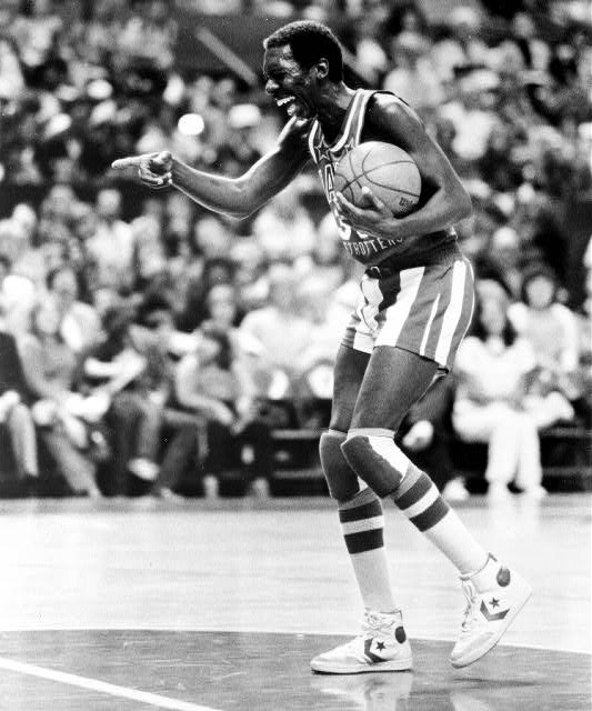 Black man in "Harlem Globetrotters" basketball uniform with ball on court smiling pointing with audience background