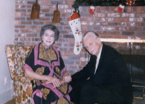 Elderly white woman in colorful dress and elderly man in suit seated holding hands by fireplace and Christmas stockings