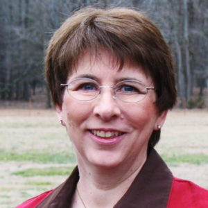 White woman with short hair and glasses smiling in red jacket and brown open collared shirt