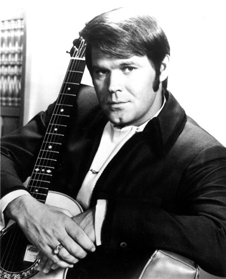 White man in suit posing with his arms around an acoustic guitar