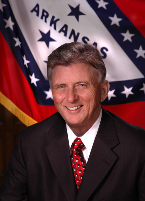 portrait white man smiling in suit and tie in front of Arkansas flag