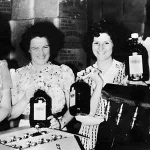 Four smiling white women in dresses in front of product boxes modeling large bottles of wine.