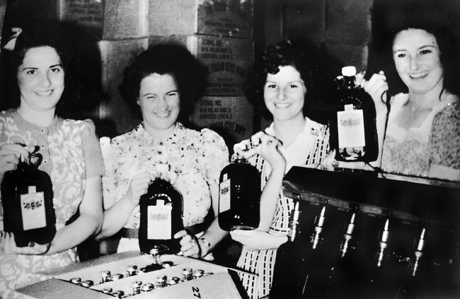 Four smiling white women in dresses in front of product boxes modeling large bottles of wine.