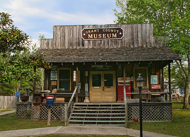 Single-story storefront building with covered porch and "Grant County Museum" sign under tree on grass