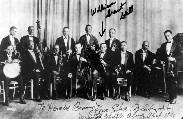 Group photo showing two rows of thirteen black men in tuxedos with instruments on stage with handwriting and arrow pointing to "William Grant Still"