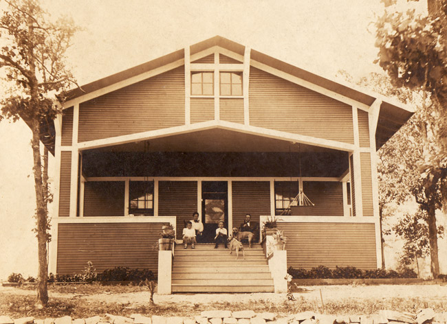 Wood frame home with man woman and two boys seated on  wide covered porch with two dogs