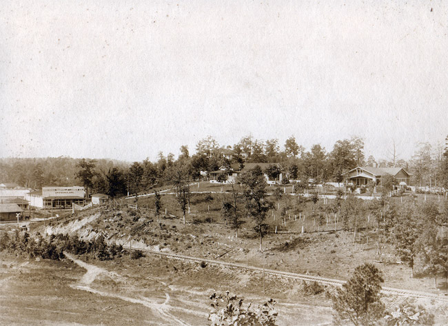 Multistory house and guest house on tree covered hill with buildings below at its base on dirt road