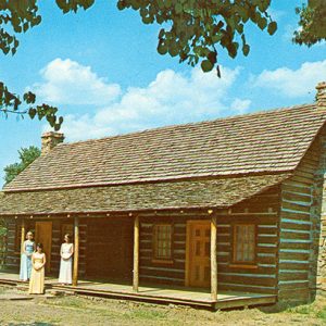 Three white women in dresses standing outside log cabin with covered porch