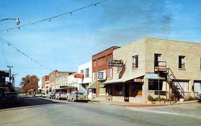 Street with brick buildings, parked cars, overhanging string lights and various business signs