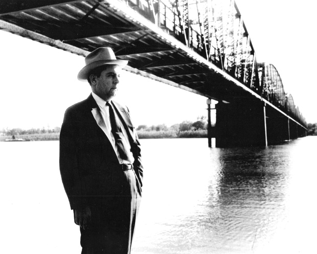 white man in suit and hat standing below bridge