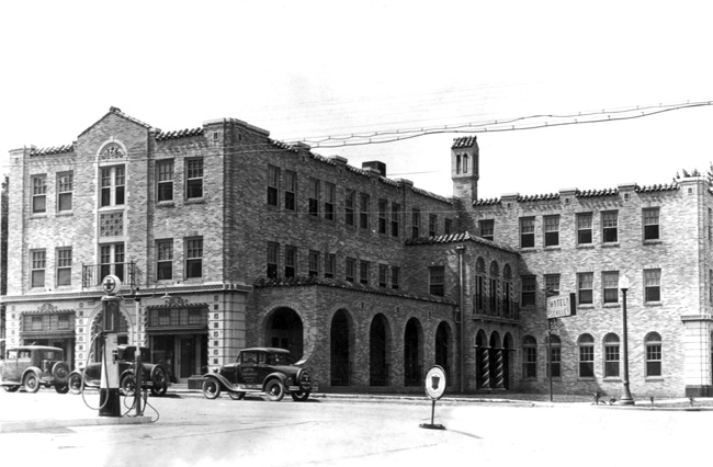Three story brick architecture with "Hotel" sign, archway tunnel, spiral columns, barrel tiles, cars, fuel pump in foreground