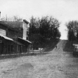 Street scene with dirt road rising over  wooded hill running alongside stores labeled "Drugs" and "Bakery" opposite hitched horses.