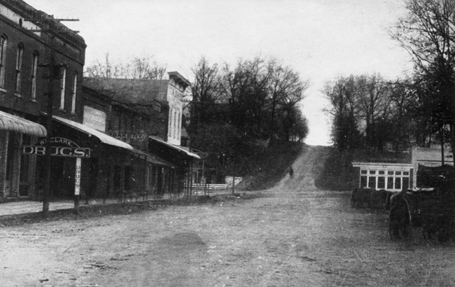 Street scene with dirt road rising over  wooded hill running alongside stores labeled "Drugs" and "Bakery" opposite hitched horses.