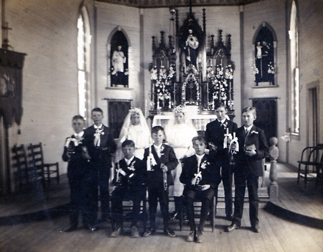 Group photo before the altar at a Catholic church, seven white boys in suits holding candles, two white girls in dresses, veils