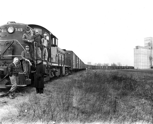 Group of white men posing on and standing by train engine with cars stretching around corner with multistory building in the background