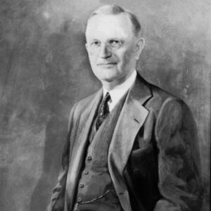 Portrait of white man standing smiling in glasses three piece suit with his hand on chair holding paper