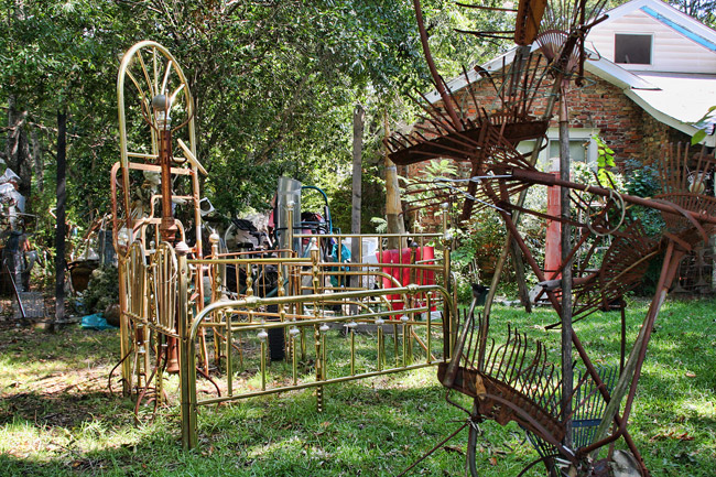 sculpture made from several brass headboards and footboards, and another made with rusted yard brooms, in front of brick house