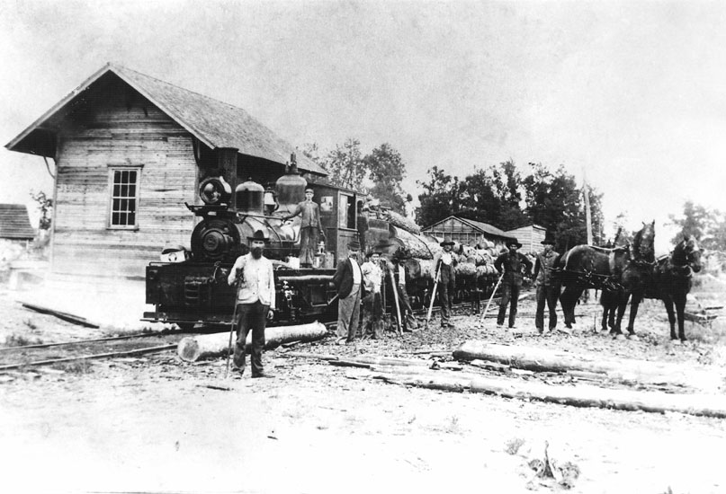White men in hats and two horses standing alongside steam locomotive still beside depot