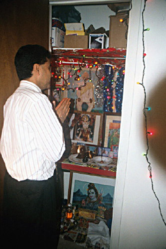 Hindu man in collared shirt slacks praying to shrine in closet with  strand lights incense