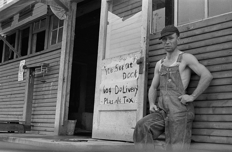 White man in cap and overalls standing outside his house with sign on open front door
