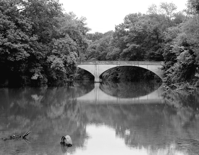 double arch support bridge over reflective forested river, half-submerged car wheel in shallow river foreground