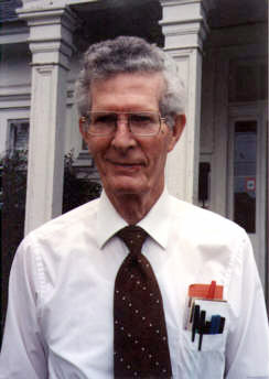 Old white man with glasses smiling in shirt and tie with pens in pocket protector