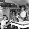Two white boys and man examine books in home library with table and window