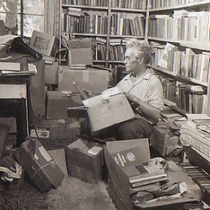 White man kneeling, holding book, among stacks of boxes in home library