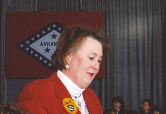 white woman in red coat in front of Arkansas state flag