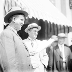 White men in suits and hats watching white man in suit and hat holding papers in his left hand standing next to white man with glasses in suit and hat