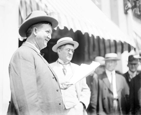 White men in suits and hats watching white man in suit and hat holding papers in his left hand standing next to white man with glasses in suit and hat