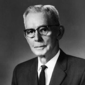 Portrait photograph of an older white man in suit tie glasses pocket square with short parted hair and a solemn expression