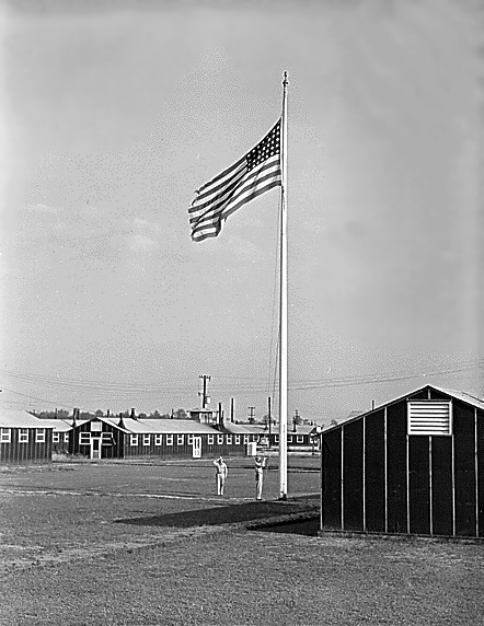 Flag raising with two soldiers, one saluting, in field near barracks.