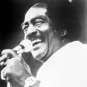 smiling Black man with receding medium hair singing into microphone wearing pinky ring and watch