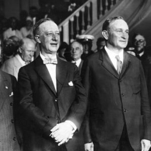 Three white men in row suits ties smiling looking to side with crowd behind