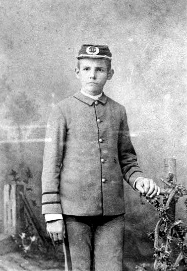Portrait of white boy in uniform with cap standing with hand on vine-covered fence post