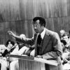 Black man in suit and tie speaking at a lectern gesturing with audience seated behind