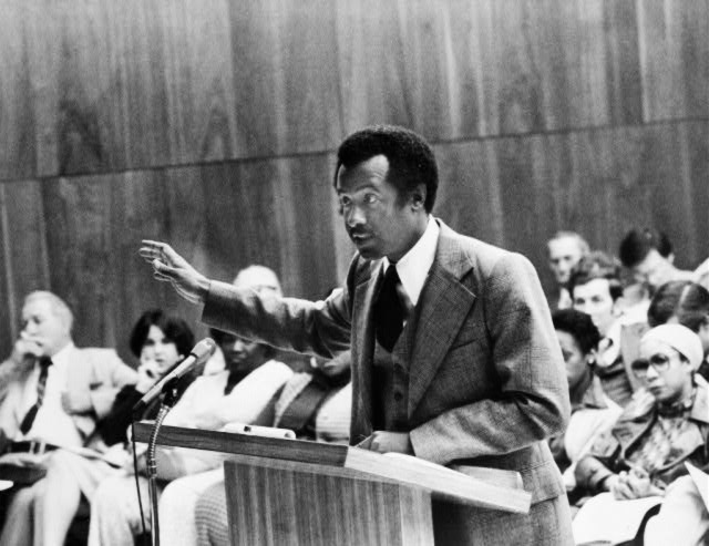 Black man in suit and tie speaking at a lectern gesturing with audience seated behind