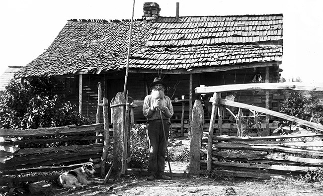 Old white man with beard standing outside cabin with covered porch and wooden fence