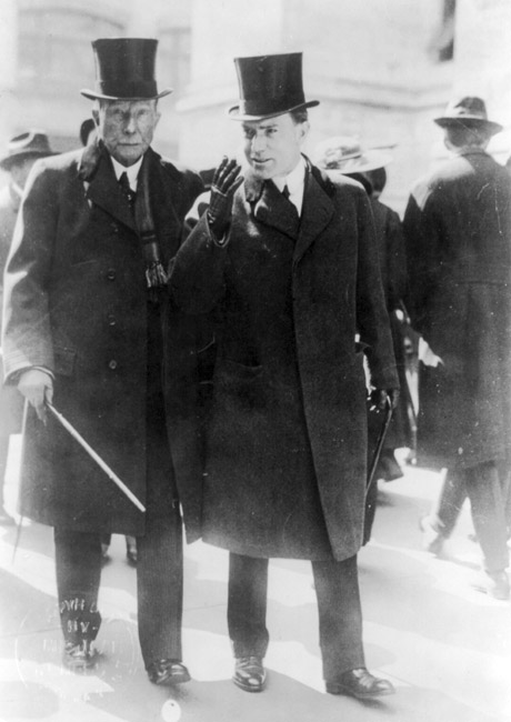 Two white men walking in suits overcoats top hats with canes on sidewalk with pedestrians