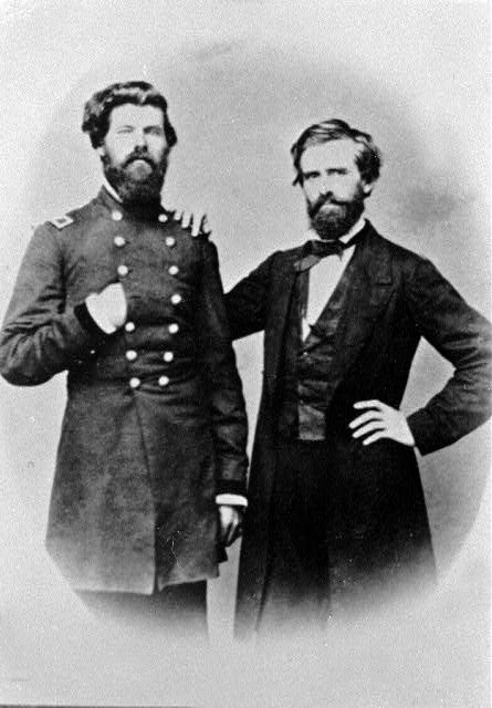 Portrait photo of a bearded white man in military uniform with another white man in a suit and bow tie with his hand on the military man's shoulder