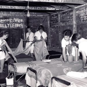 Young white women making their beds in cabin dormitory with names written on the walls around them