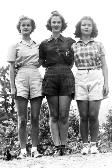 Three young white women in shirts and shorts standing with their arms around each other's backs