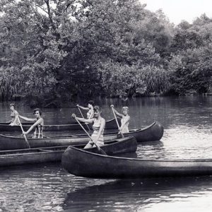 Young white women paddling in canoes on lake with trees in the background