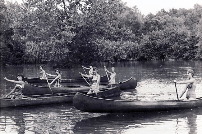 Young white women paddling in canoes on lake with trees in the background