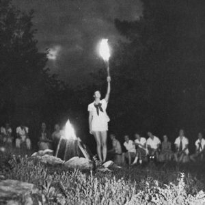 Young white women sitting around a campfire watching a young white woman in a shirt and shorts holding up a lit torch
