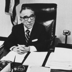 Older white man suit glasses hands crossed at cluttered office desk U.S. flag and ashtray behind