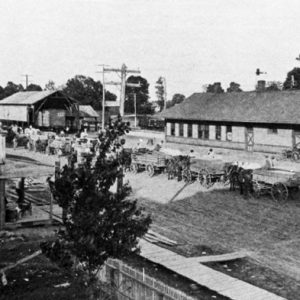 Horse and carriage procession along train depot with building sign reading "Mary Jane Table Syrup"