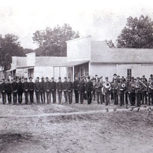 military group photo, with band members specifically numbered 1 to 8, located on dirt road lined with wood framed buildings